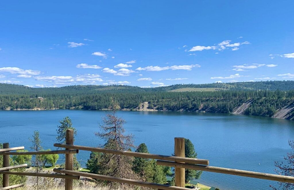A wide view of a vast, calm lake and forested mountains seen over a rustic wooden log fence on a bright sunny day, representing boundaries and the open possibilities of life after a crisis.