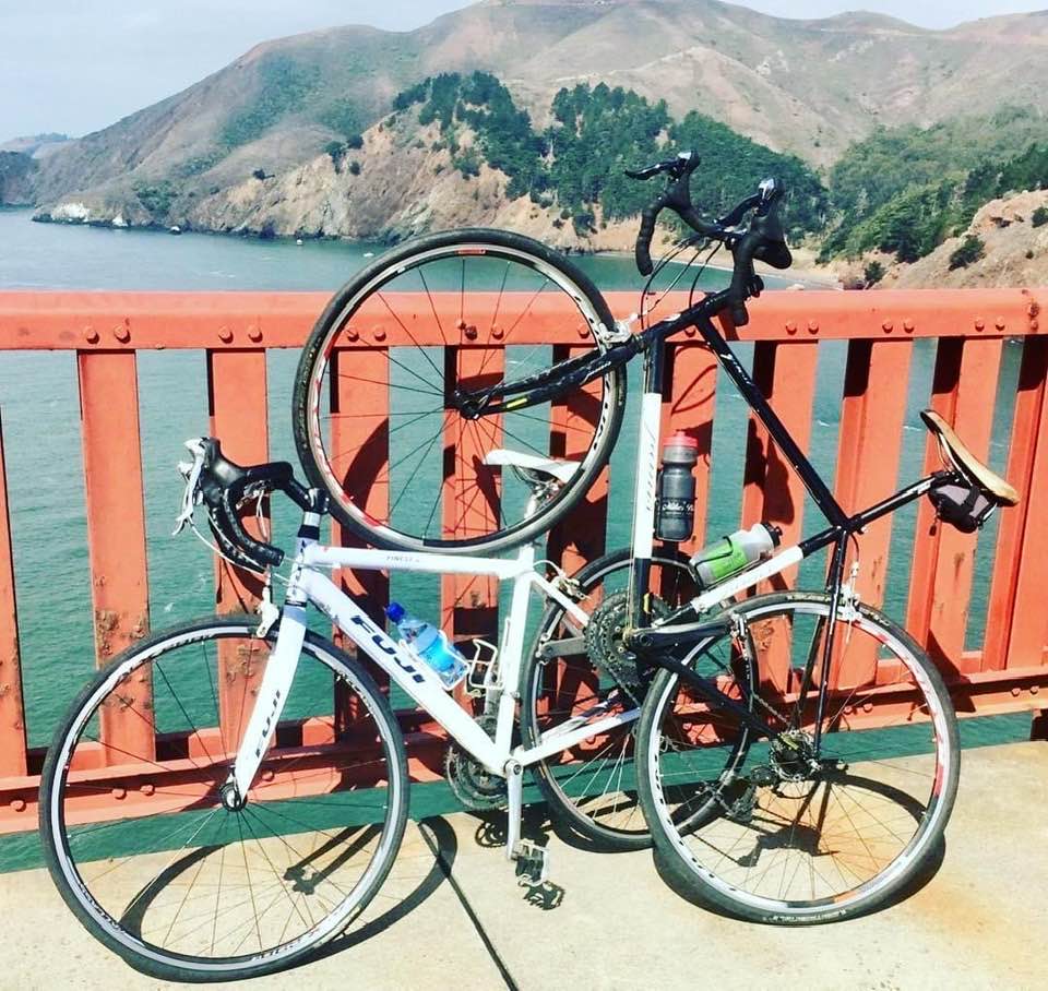 Two road bicycles, a white Fuji and a black frame with Fuji components, propped precariously on the Golden Gate Bridge railing, with the Pacific Ocean and Marin Headlands in the background. The unique pose of the black bike's elevated front wheel symbolizes freedom and intentional movement.