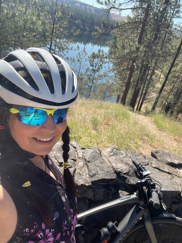 Donna aging well, smiling on a bike ride with helmet and sunglasses, lake and trail in the background